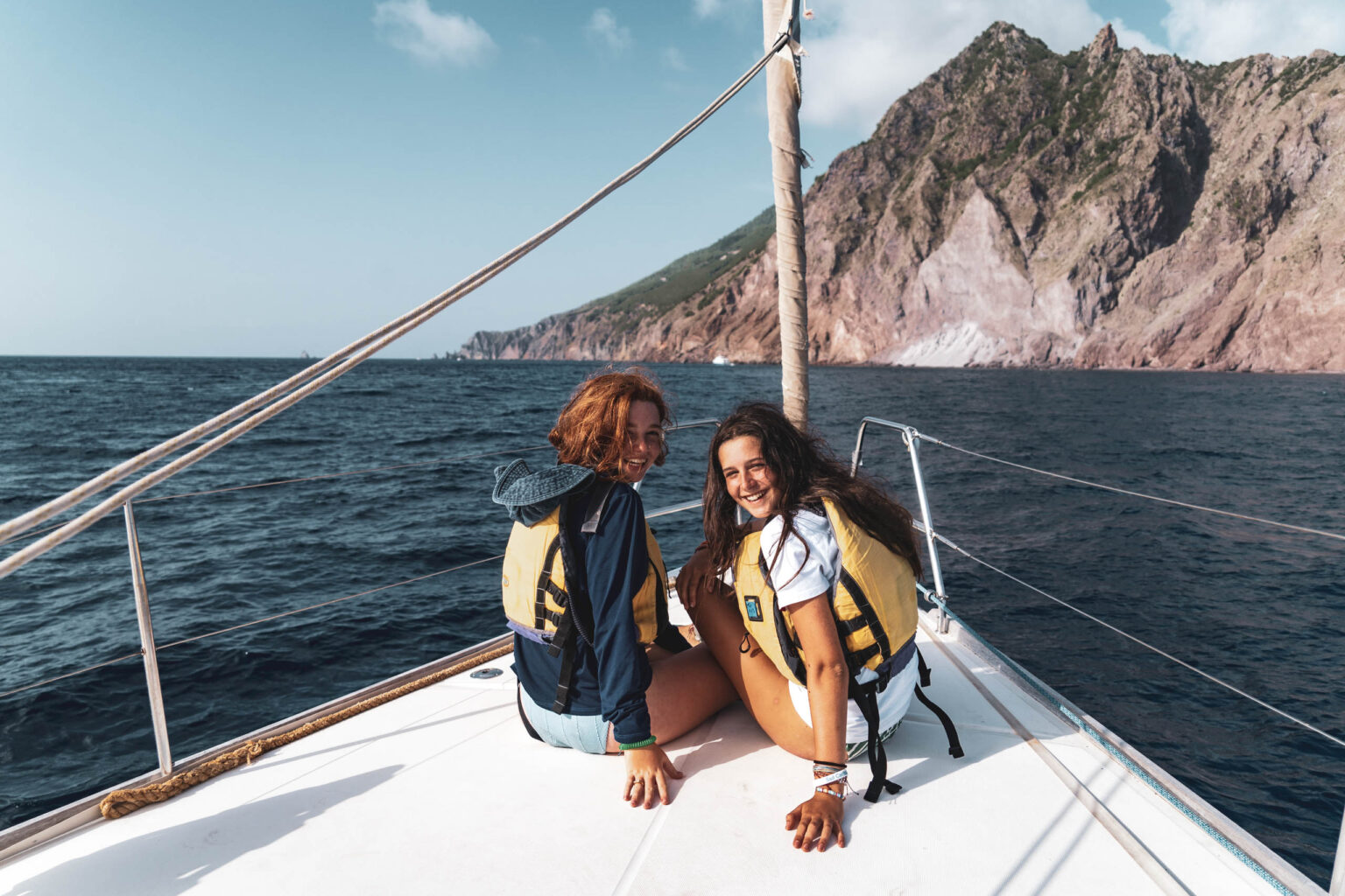 Two women sitting on the back of a sailboat in the ocean.