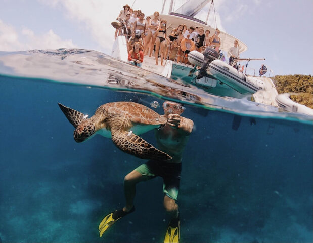 A man swimming with a turtle under water with people in the background.