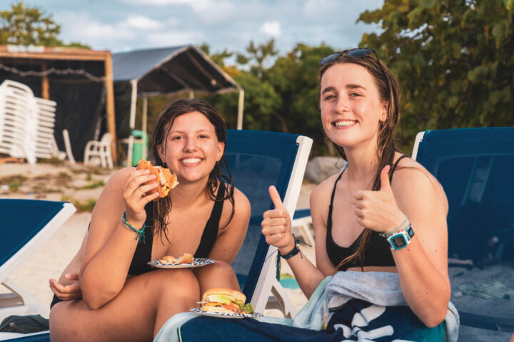 Two campers sitting on the beach eating a hamburger.