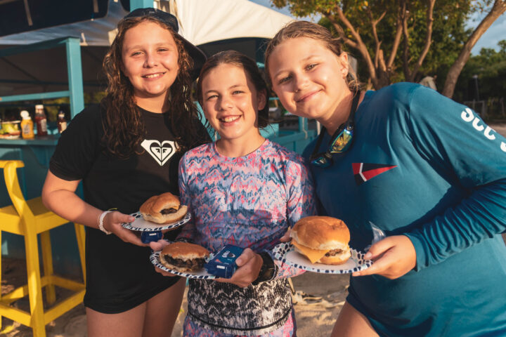 Three girls holding plates of hamburgers on the beach.