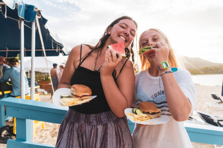 Two women eating burgers and watermelon on the beach.