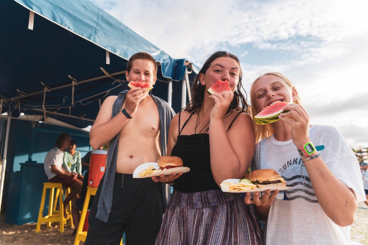 Campers eating watermelon and hamburgers at a beach.