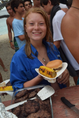 A girl wearing a blue shirt and holding a plate with a hamburger and corn on the cob.