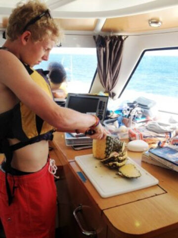 A camper in a life jacket cutting a pineapple on a boat.