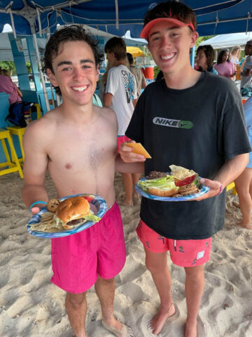 Two young men holding plates of food on the beach.