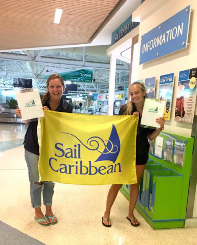 Two people holding up a Sail Caribbean flag at the airport.