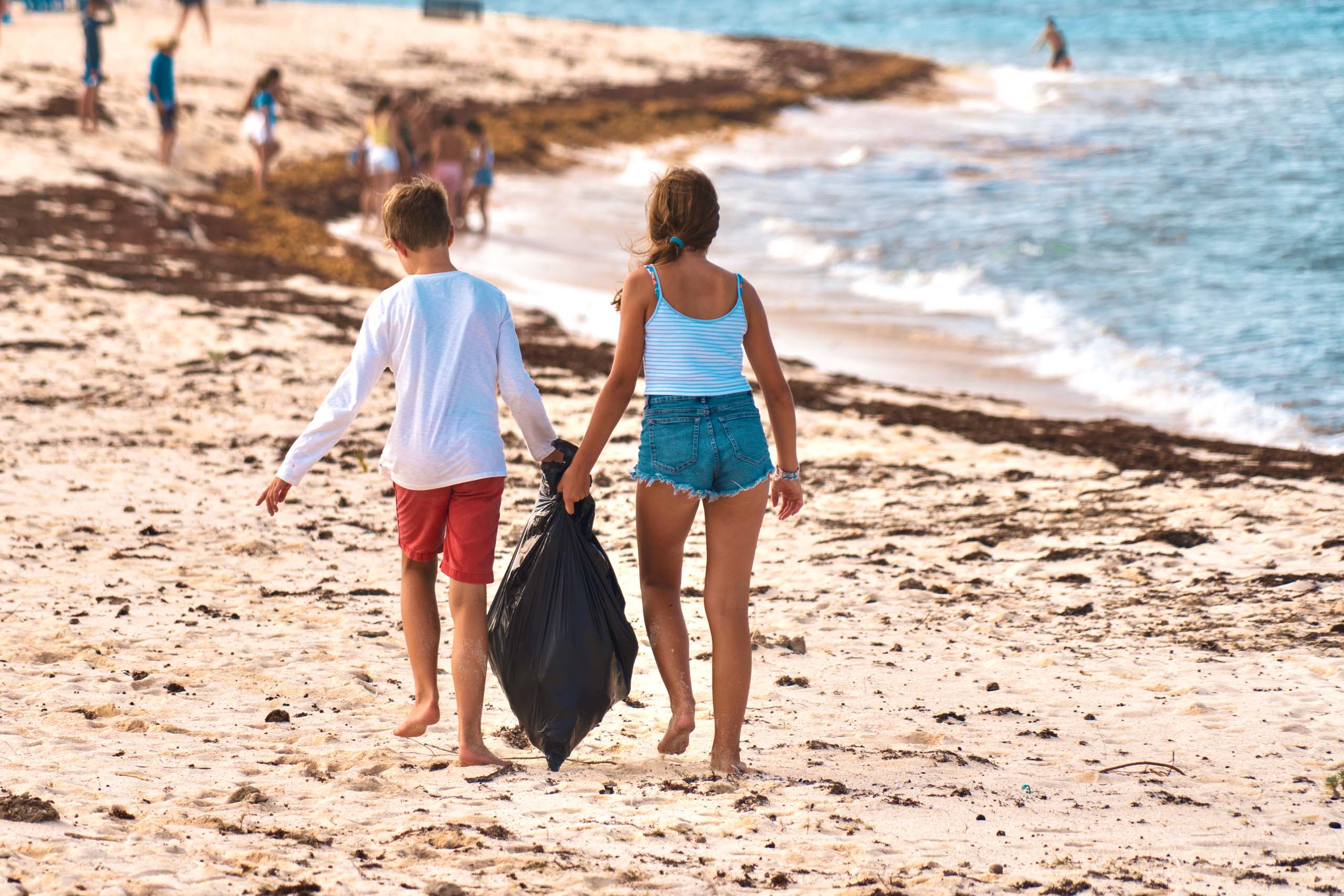 Two children walking along a beach with a trash bag.